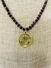 Fleur-de-lis round gold plated pendant hangs from a faceted garnet necklace